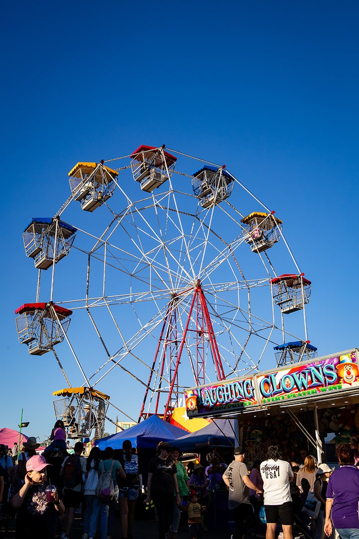 A ferris wheel. In front is a 'laughing clowns' stall. There is a large crowd of people.
