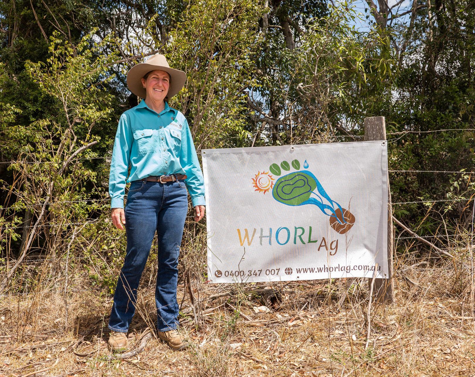 Marcia stands in front of a Whorl Ag sign hanging from a wire fence. The fence is in a brown paddock against bushy trees.