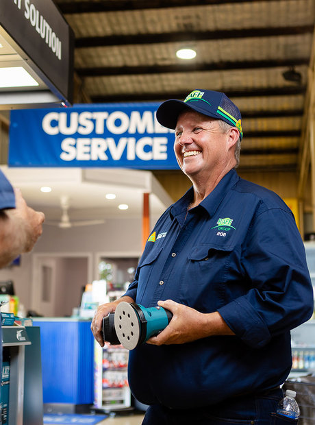 A RAFF Group employee holds an electric sander while talking to a customer. In the background you can see a 'customer service' banner.