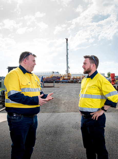 Two men wearing hi-viz jackets are talking on a driveway into an equipment yard