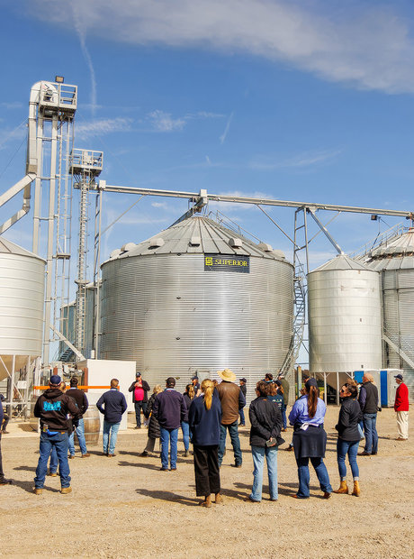 A large group of people gather around at the Alvan Blanch yards. There are large feed silos in the background. 