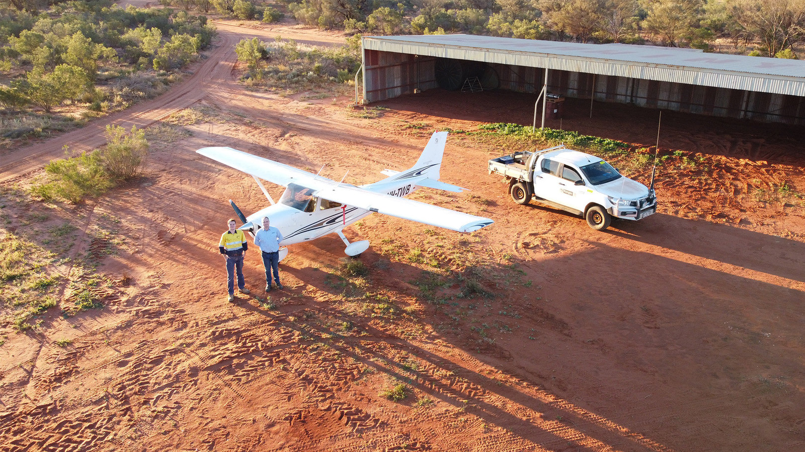 Two men stand next to a small plane on a dirt runway. To the right behind them is a ute and a large empty shed,