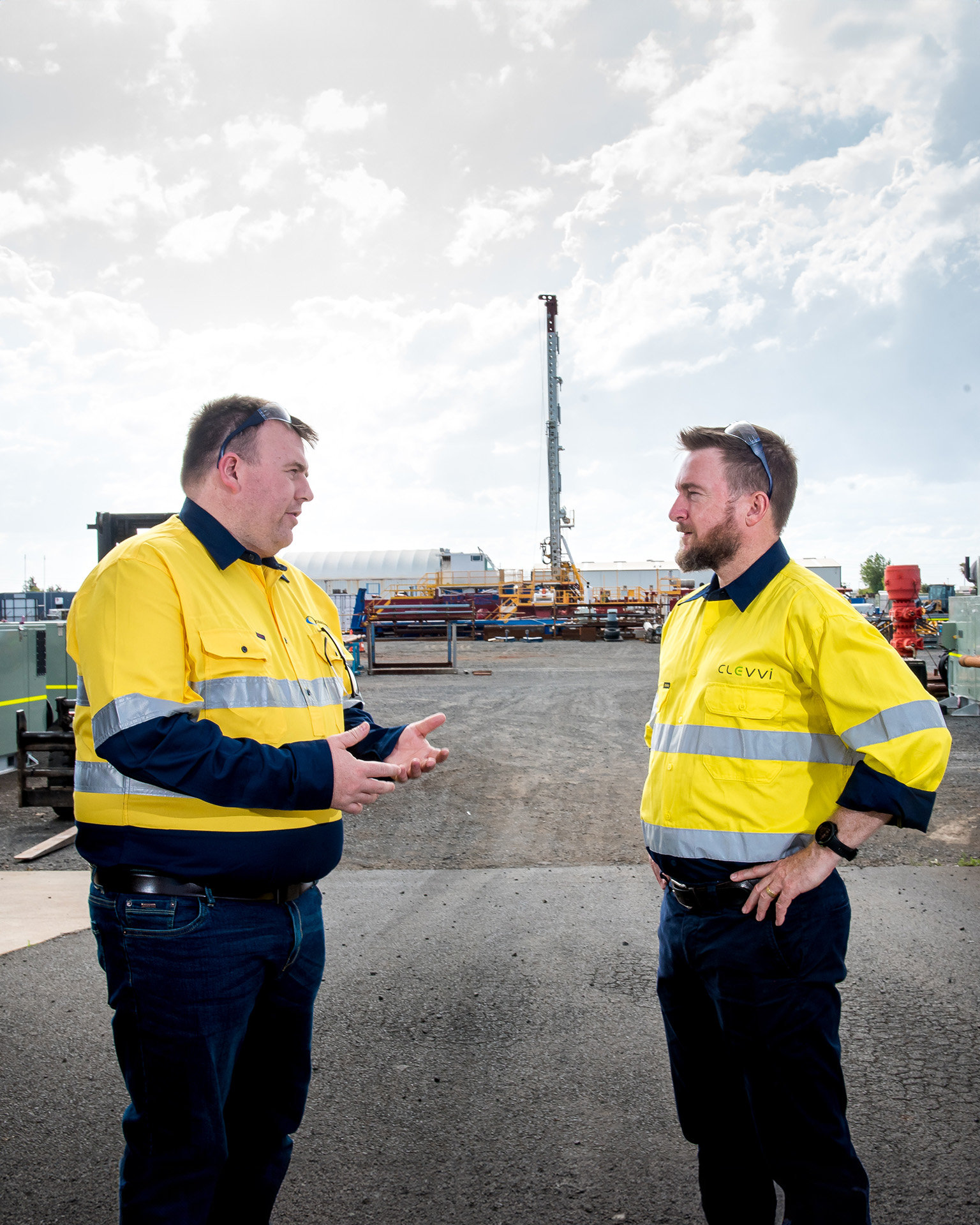 Two men wearing hi-viz jackets are talking on a driveway into an equipment yard