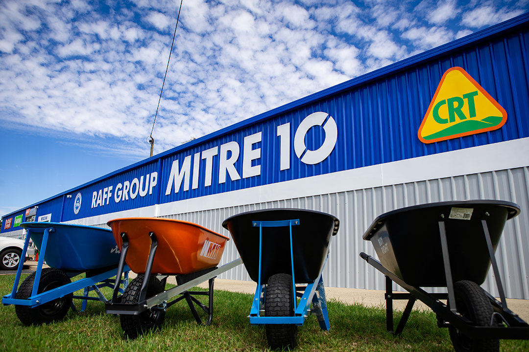 4 different coloured wheelbarrows sit on a lawn outside a RAFF Group Mitre 10 building.